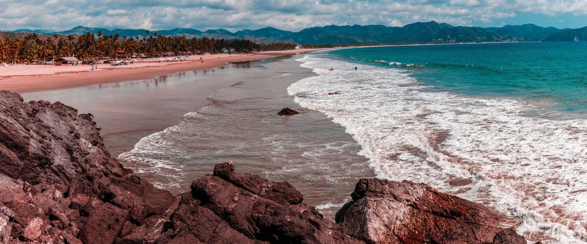 A beach with rocky foreground, a sandy shore, and a mountain range in the background.