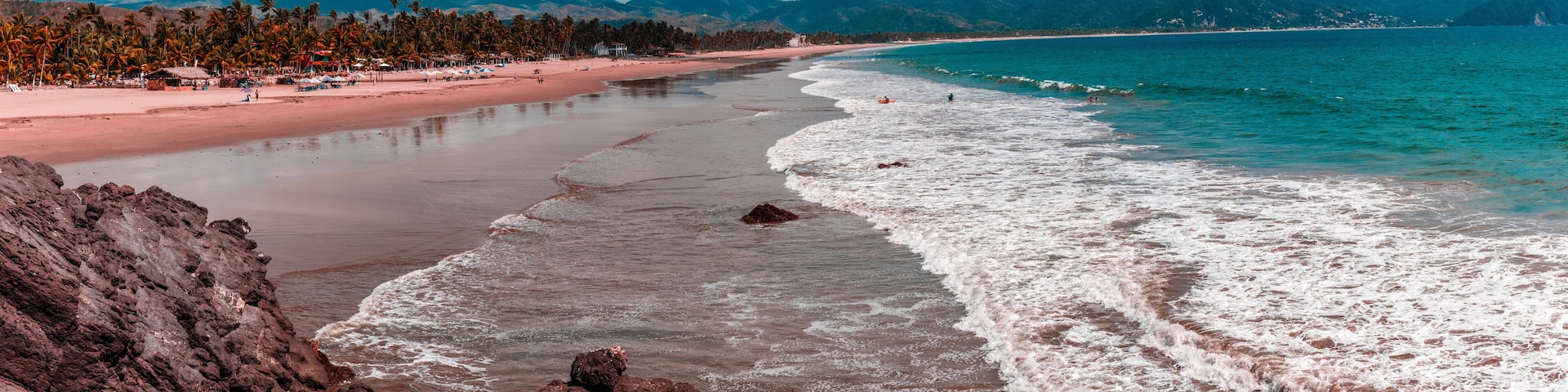 A beach with rocky foreground, a sandy shore, and a mountain range in the background.