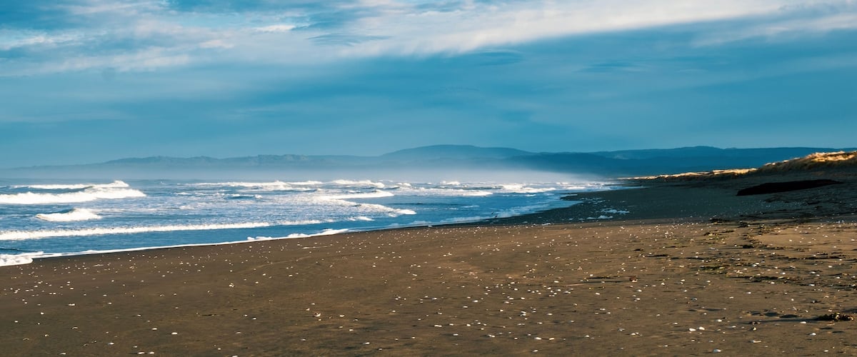 A beach with waves, a clear sky, and distant mountains.