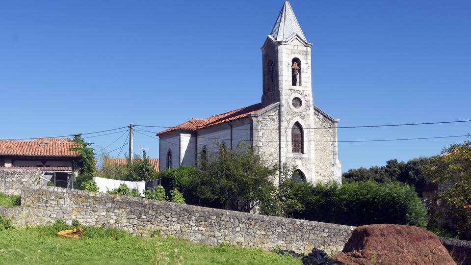 A stone church with a bell tower and a red-tiled roof, surrounded by greenery and a stone wall.