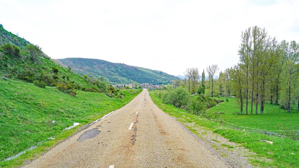 A rural road flanked by green hills and trees, leading towards a distant village.