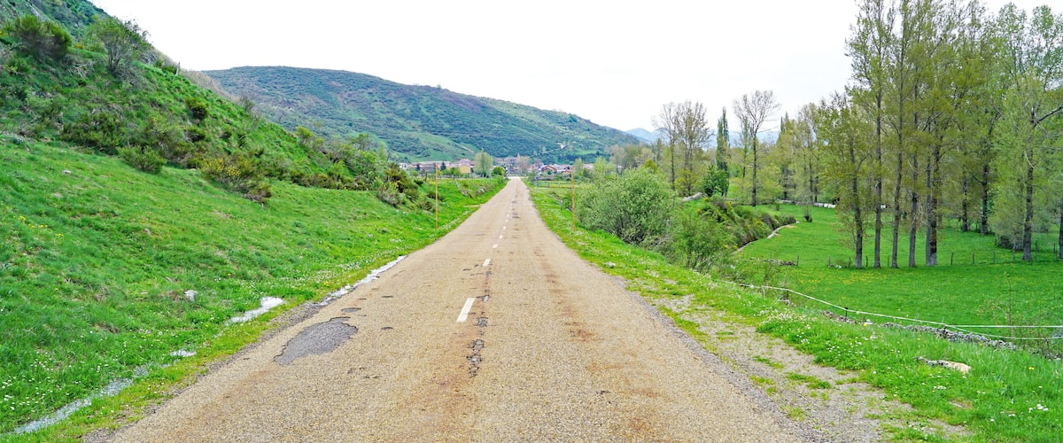 A rural road flanked by green hills and trees, leading towards a distant village.