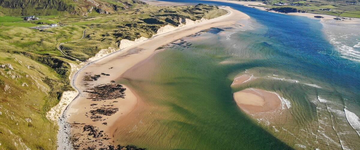 A coastal landscape with a sandy beach, green hills, and a clear blue sea.