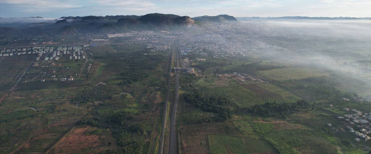 An aerial view of a city with a prominent highway, green fields, and a mountain in the background.