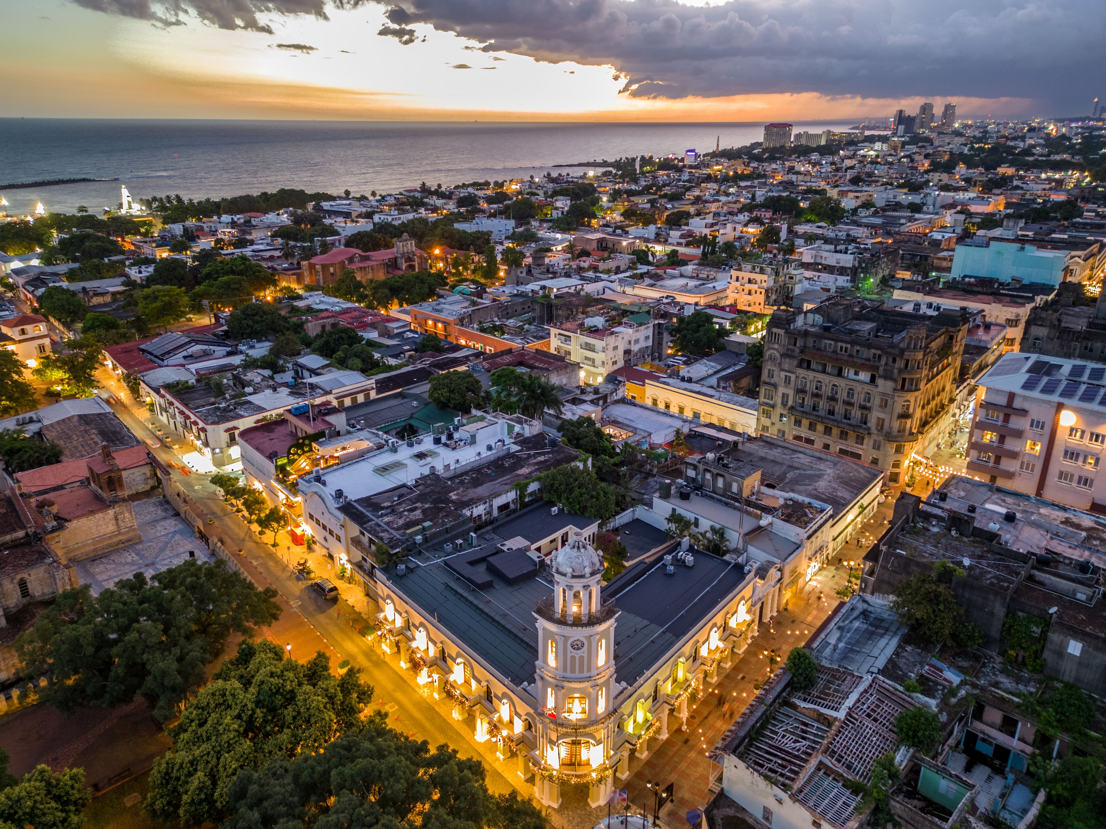 Paisagem costeira ao crepúsculo com edifícios iluminados, uma igreja em destaque e vista para o mar.