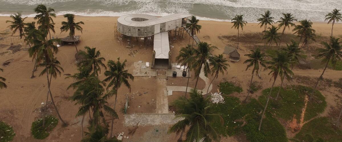 A circular building with a spiral staircase on a beach with palm trees.