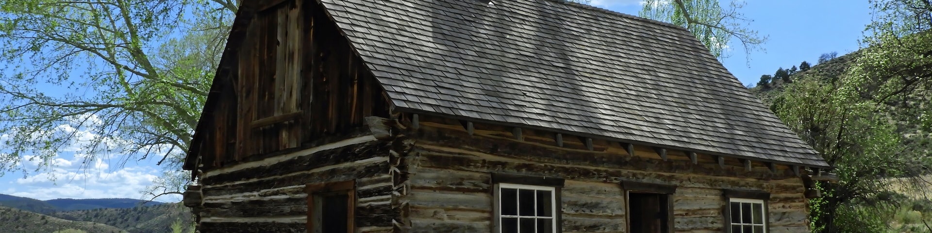 A rustic log cabin with a steep gabled roof and a chimney, surrounded by a rural landscape with rolling hills and a clear blue sky.