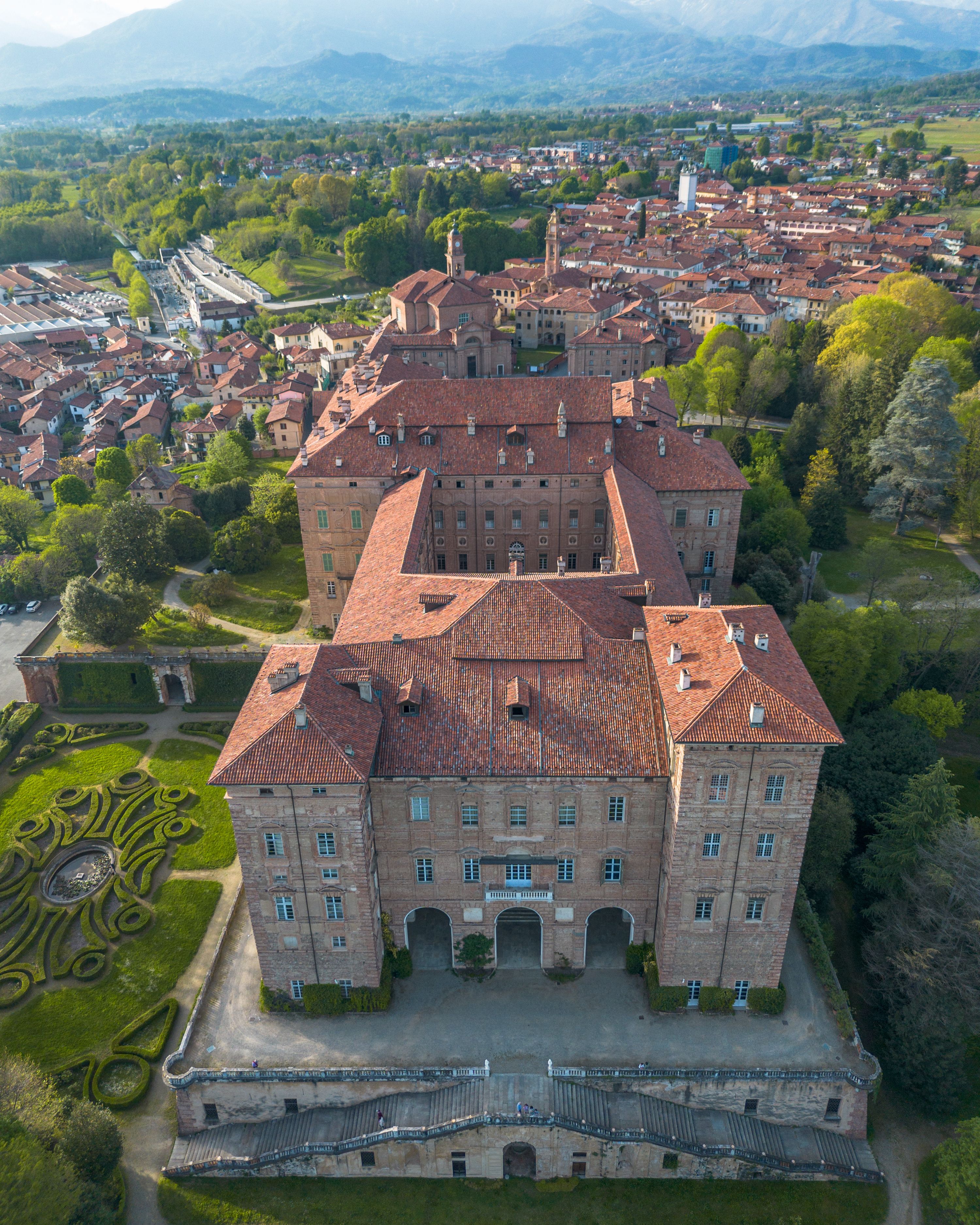 A large, historic building with a red-tiled roof, surrounded by lush greenery and a well-maintained garden.