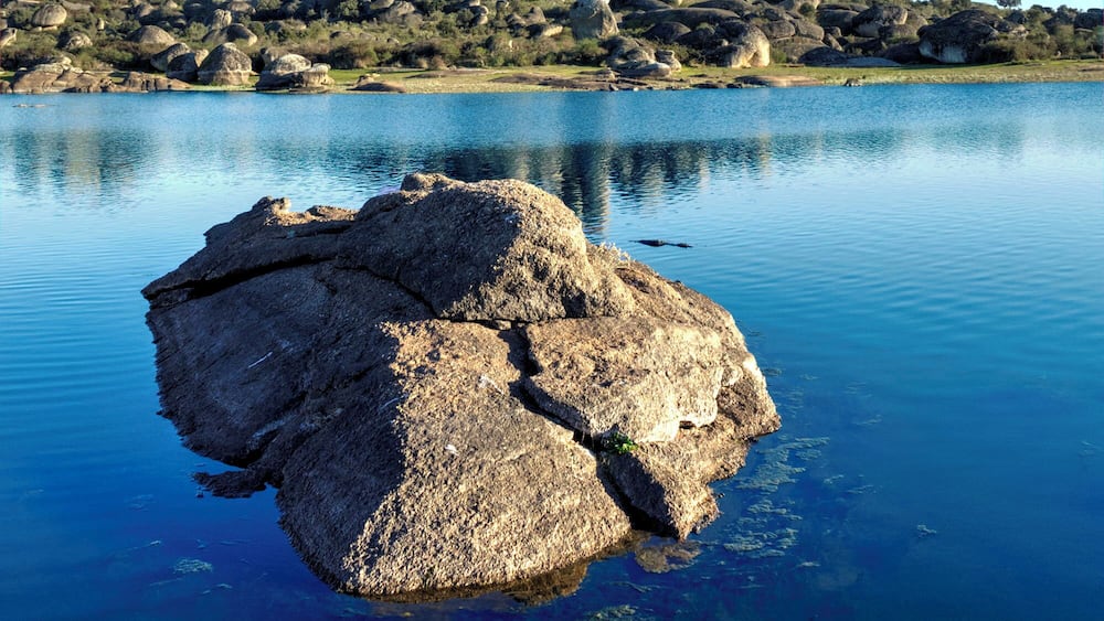 Ein ruhiger See mit einem markanten Felsen im Vordergrund und einer felsigen Hügelkette im Hintergrund.