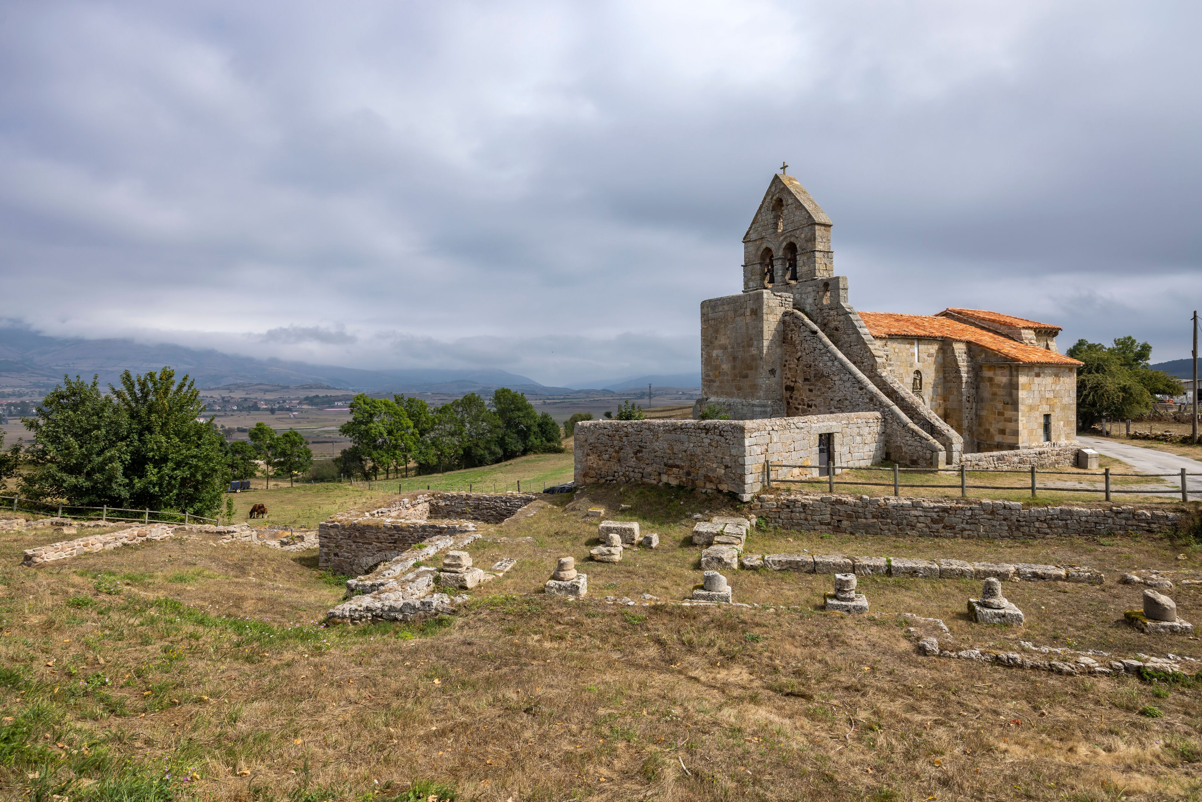 An ancient stone church with a bell tower, surrounded by ruins and a rural landscape.