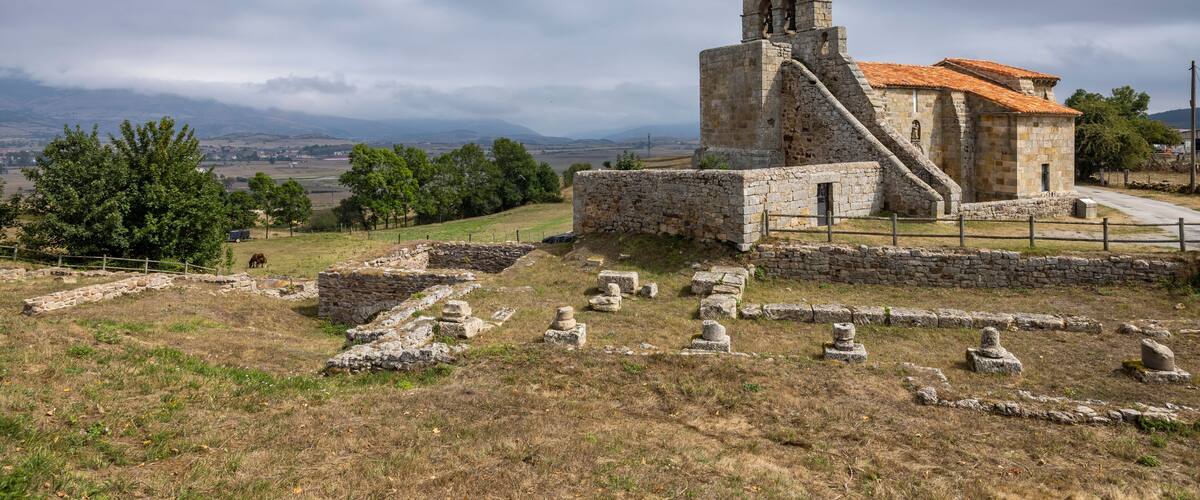 An ancient stone church with a bell tower, surrounded by ruins and a rural landscape.