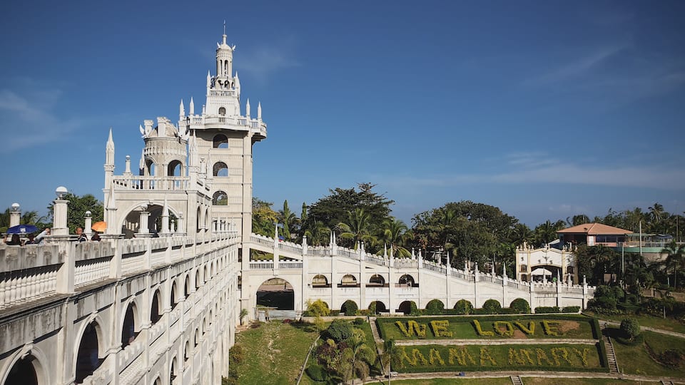 A grand white castle with a tall tower, arched walkways, and a garden with 'We Love Maria' written in flowers.