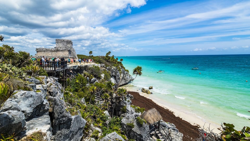 A coastal view with a clear blue sky, turquoise sea, and a stone structure with visitors.