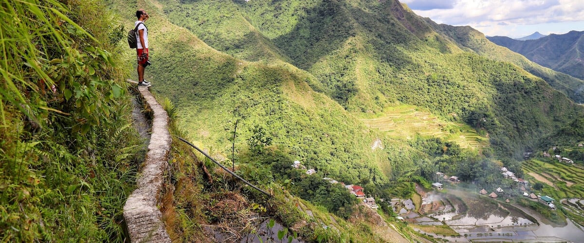 A hiker on a narrow path with terraced fields and mountains in the background.