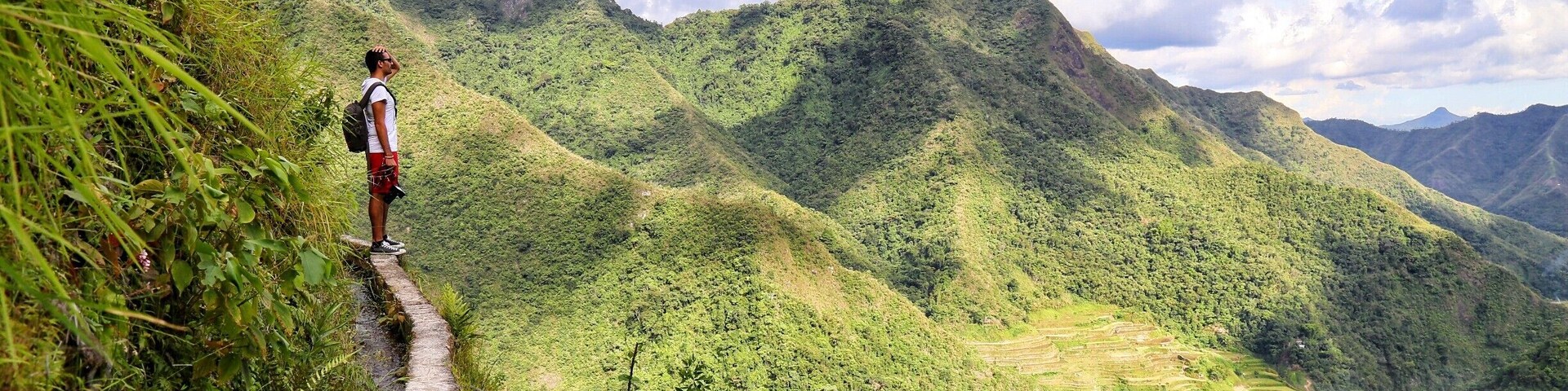 A hiker on a narrow path with terraced fields and mountains in the background.