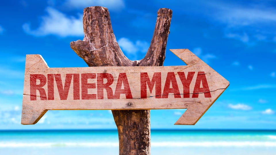 A wooden sign with 'RIVIERA MAYA' written on it, pointing to the right, set against a beach with clear blue water and a blue sky.