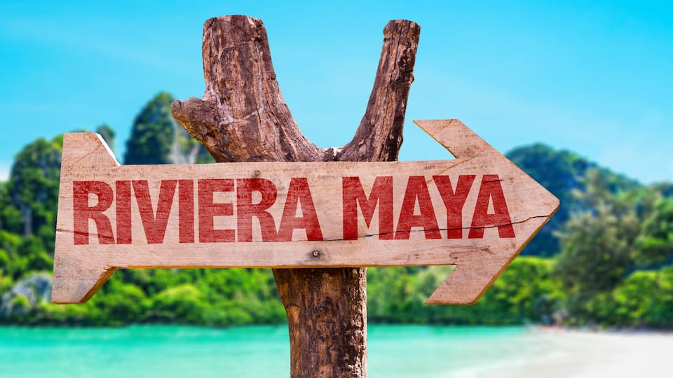 A wooden sign with 'RIVIERA MAYA' written on it, pointing to the right, with a beach and clear blue sky in the background.