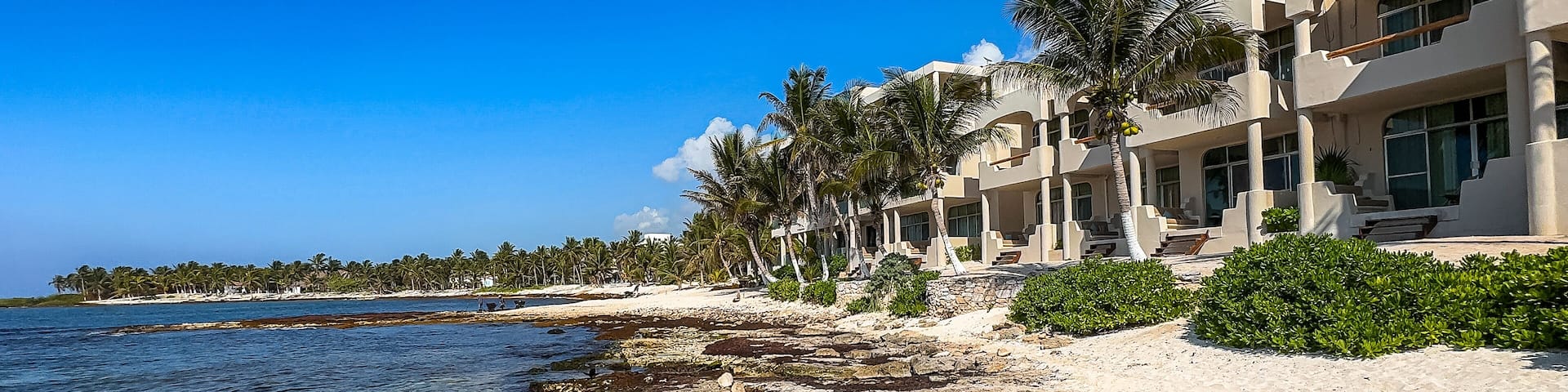 A beach with a rocky shoreline and a clear blue sky. The beach is empty and the houses are white