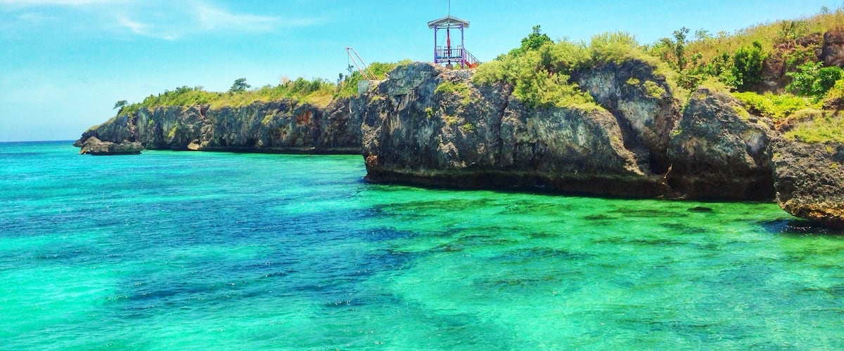 A coastal landscape with clear turquoise waters, rocky cliffs, and a small structure on top.