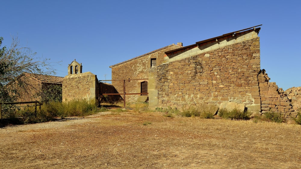 An old stone building with a bell tower, surrounded by dry grass and shrubs.