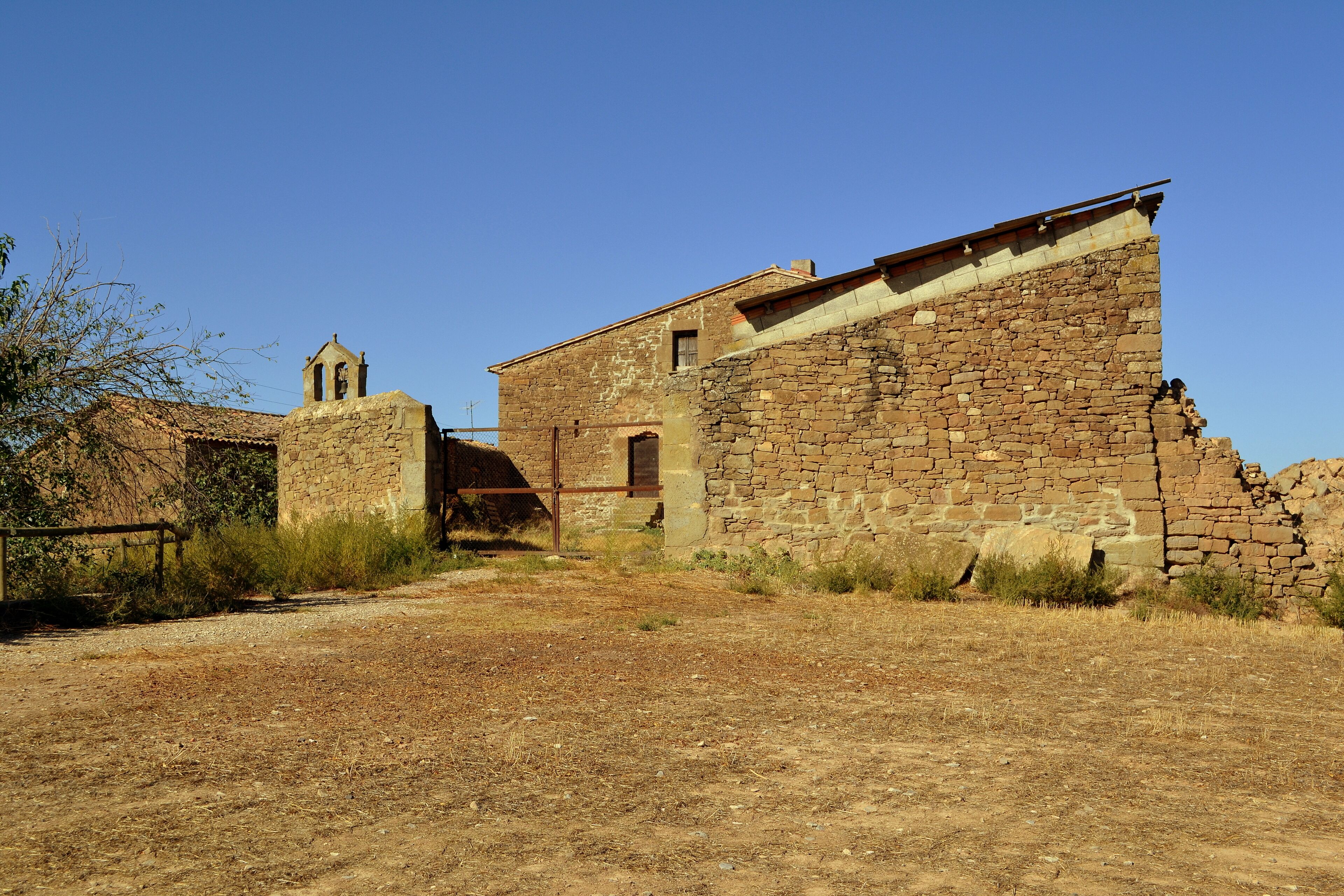 An old stone building with a bell tower, surrounded by dry grass and shrubs.