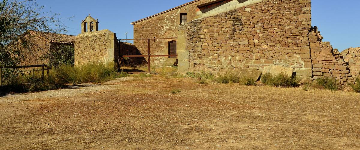 An old stone building with a bell tower, surrounded by dry grass and shrubs.