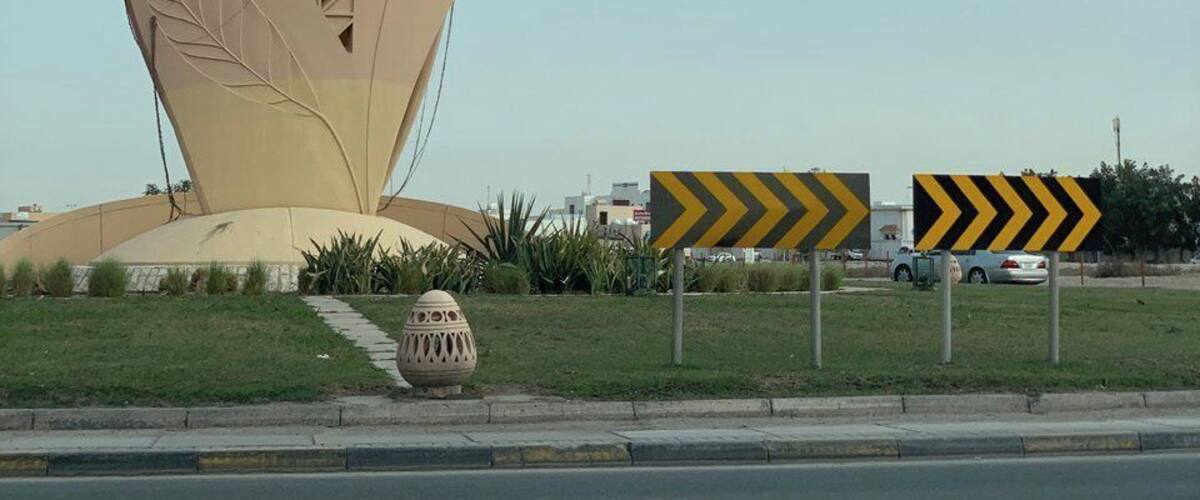 A large, abstract sculpture resembling a fish with circular eyes and a leaf pattern, located beside a road with chevron signs.