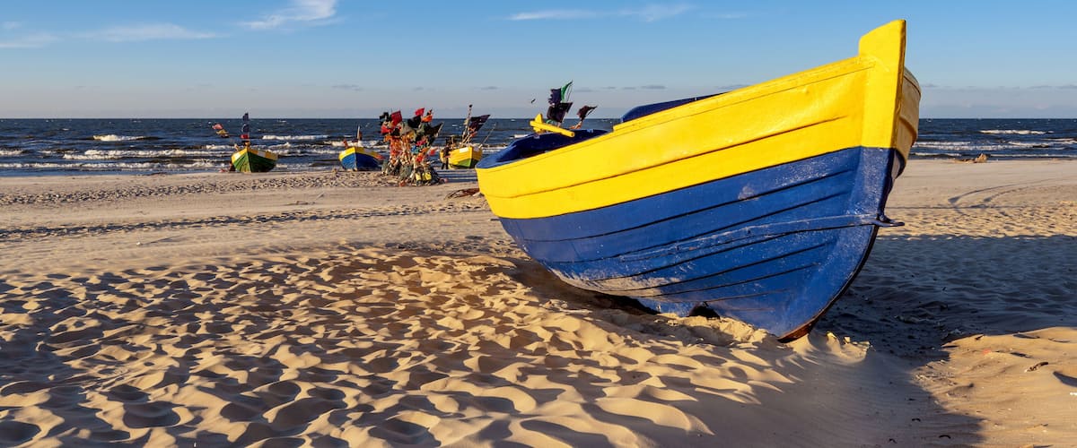 A beach with a yellow and blue boat, sand dunes, and the ocean in the background.