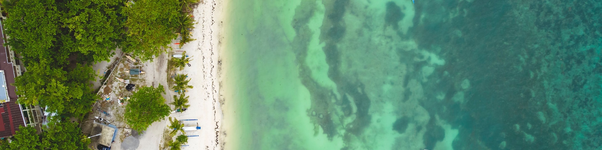 An aerial view of a beach with clear turquoise waters, a few boats, and dense green foliage.