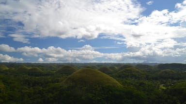 Paisagem com colinas onduladas cobertas por vegetação verde densa e céu azul com nuvens esparsas.