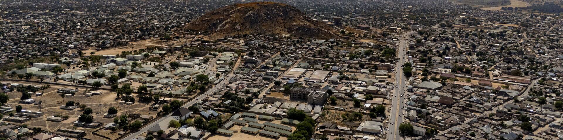 Aerial view of a sprawling cityscape nestled beneath a majestic mountain range, roads stretching like arteries through the urban landscape, Jalingo, Taraba, Nigeria.