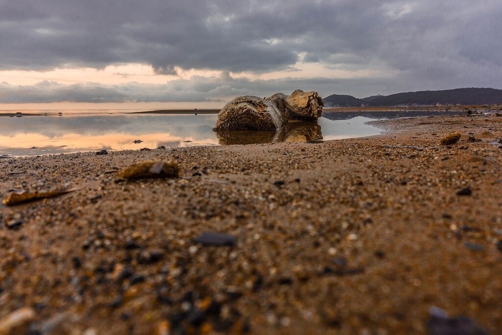 A rocky outcrop on a pebbly beach with a calm body of water and distant hills under a cloudy sky.