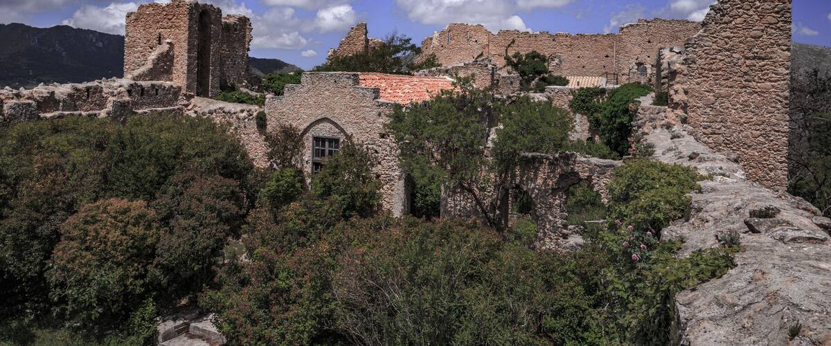 Ancient stone ruins with a red-tiled roof amidst lush greenery and mountains in the background.