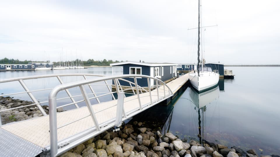 A dock with a metal walkway leading to a boat, calm water, and a small building in the background.