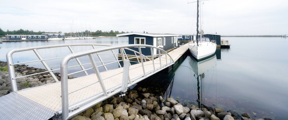 A dock with a metal walkway leading to a boat, calm water, and a small building in the background.