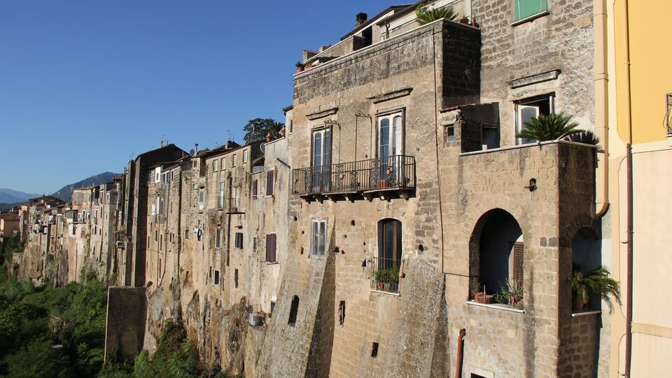 A row of historic stone buildings with balconies and arched windows, set against a clear blue sky.
