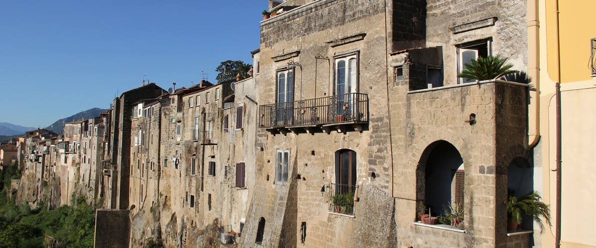 A row of historic stone buildings with balconies and arched windows, set against a clear blue sky.