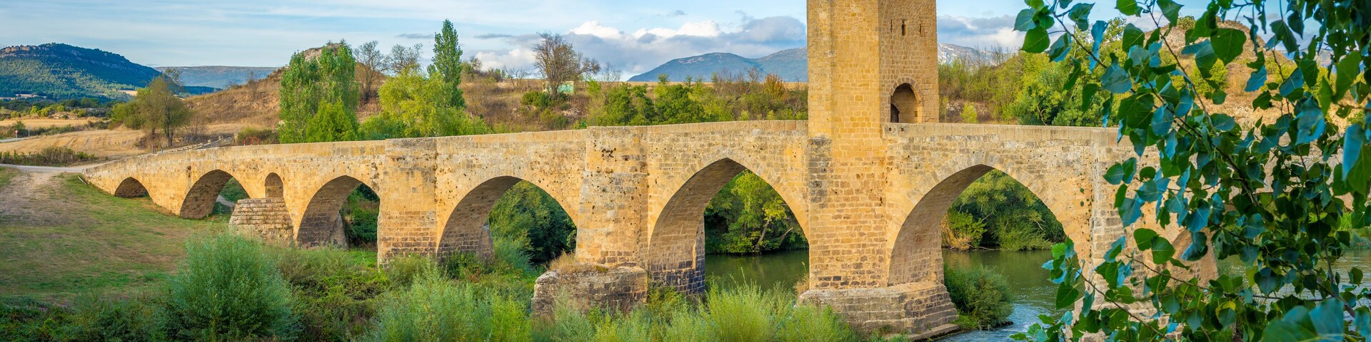 Un puente de piedra con varios arcos sobre un río, rodeado de vegetación y colinas.