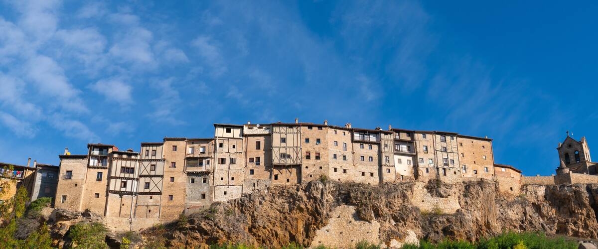A historic stone building with multiple levels and balconies, situated on a rocky cliff under a clear blue sky.