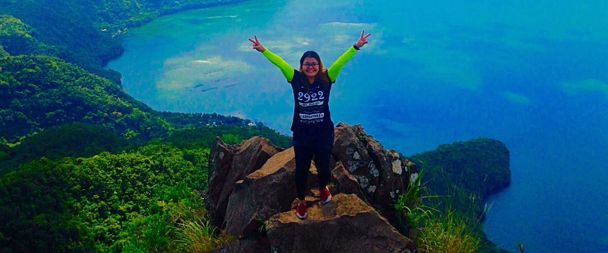 A person standing on a rocky outcrop with arms raised, overlooking a vast landscape with a large body of water and distant mountains.