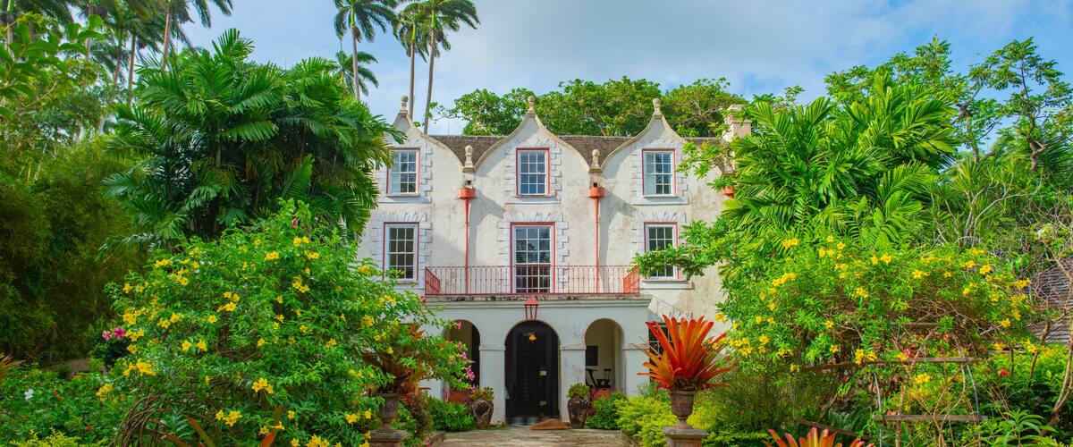 A colonial-style house surrounded by lush tropical vegetation and a paved pathway leading to the entrance.