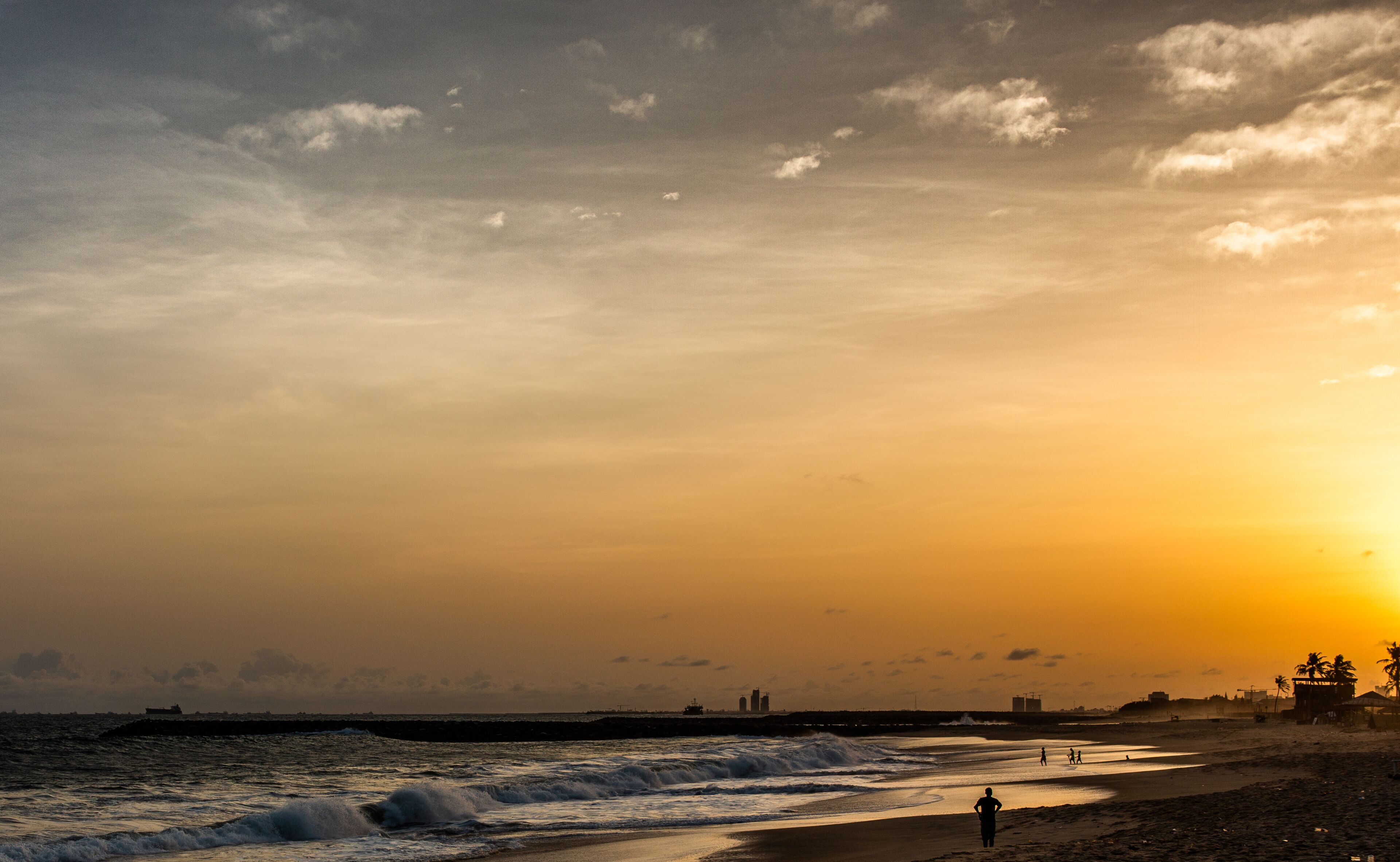 A beach at sunset with waves, a pier, and distant buildings.