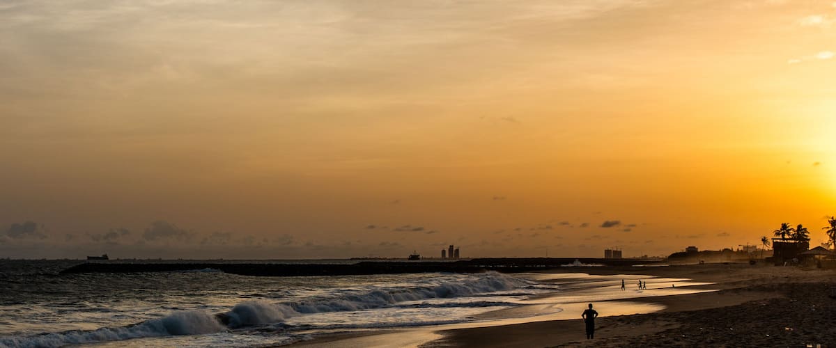 A beach at sunset with waves, a pier, and distant buildings.
