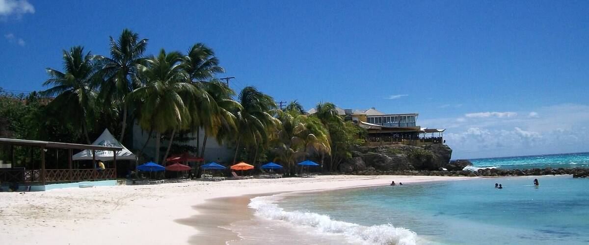 A beach with clear turquoise water, palm trees, and a building on a cliff.