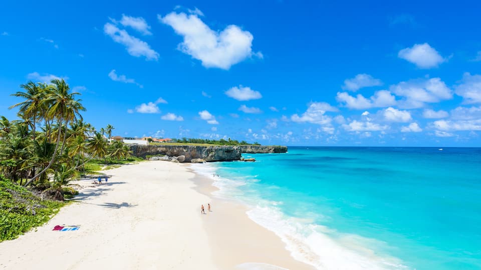 A beach with clear turquoise water, palm trees, and a rocky outcrop in the distance.