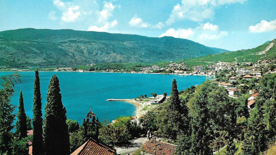 A coastal town with a large lake, surrounded by green hills and cypress trees.