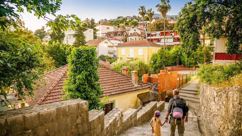A cobblestone street with a man and a child walking, surrounded by greenery and buildings.