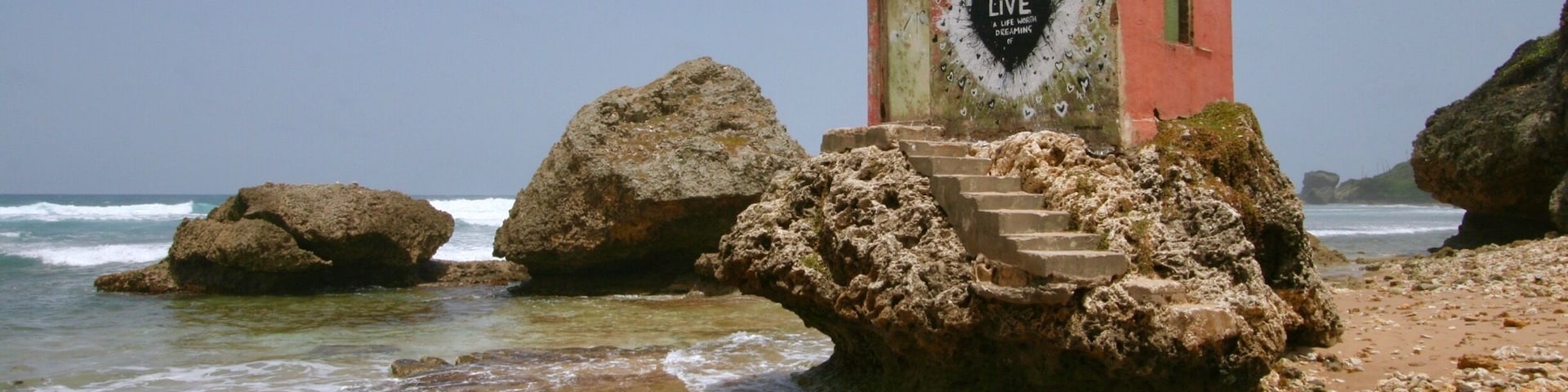 A rocky beach with a small red building featuring a heart-shaped sign that reads "LIVE A LIFE LIKE DREAMING."