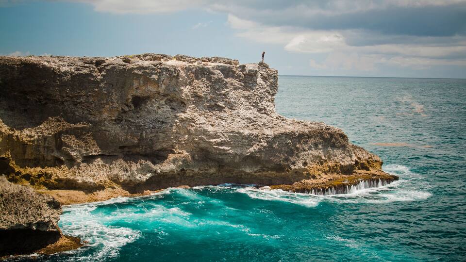 A rocky coastline with a person standing on a cliff overlooking turquoise waters.