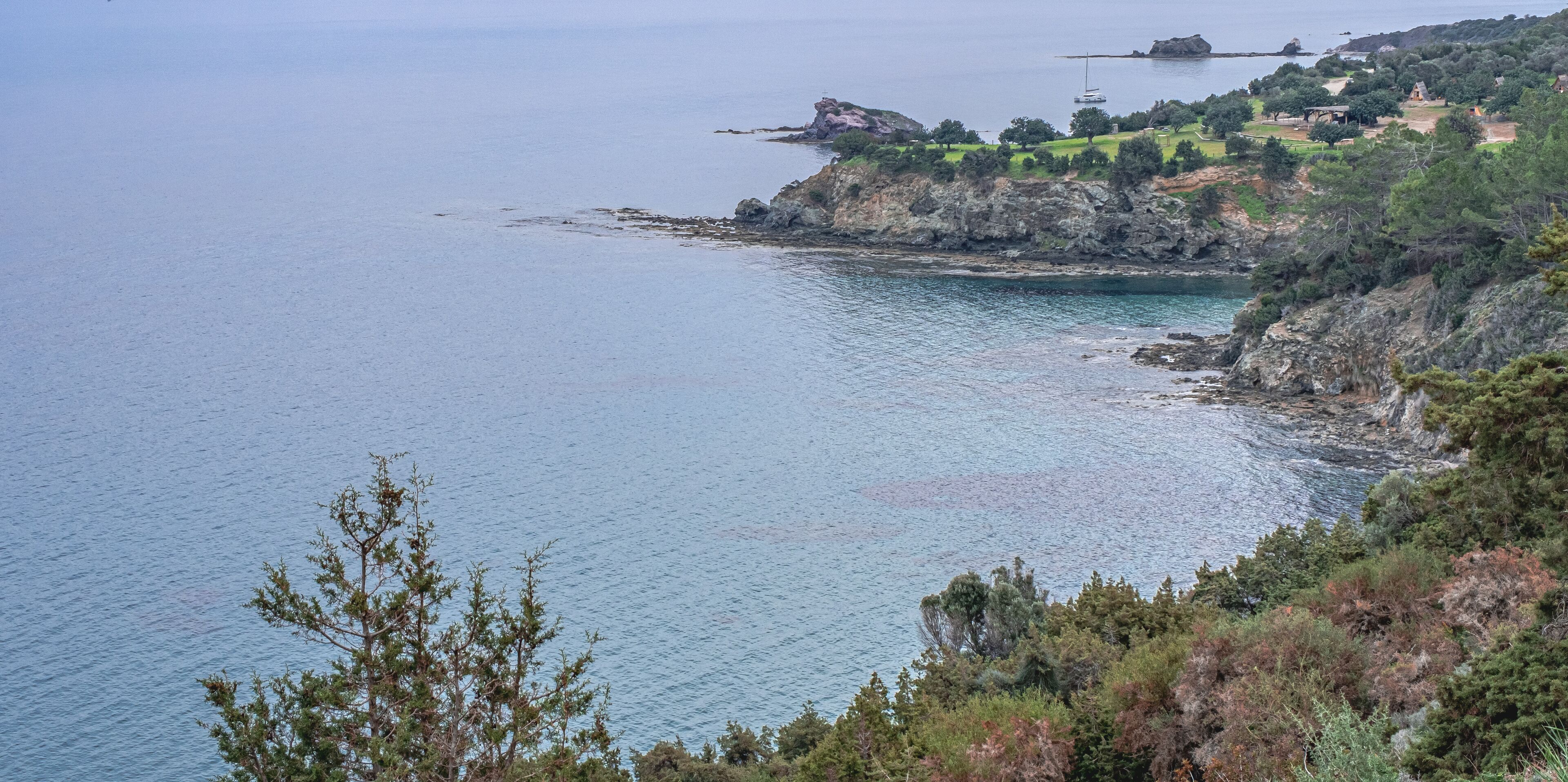 A coastal landscape with rocky cliffs, calm sea, and a small settlement with buildings and greenery.